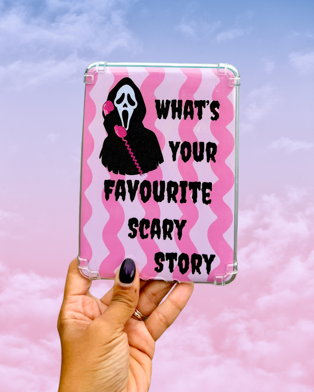 A hand with dark nail polish holds a clear-edged tablet case featuring a pink and white wavy pattern, a black and white Ghostface graphic, and the text 'WHAT'S YOUR FAVOURITE SCARY STORY' against a pink sky background.