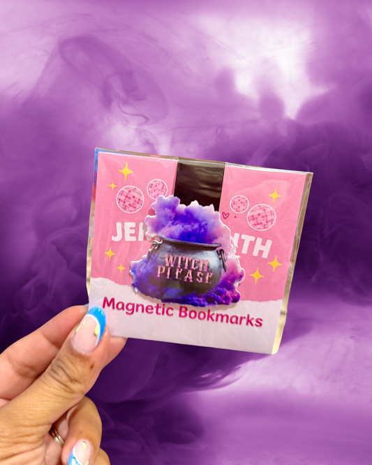 A hand holds a package of 'Witch Please' magnetic bookmarks against a purple smoky background. The bookmarks come in a pink and white package with an illustration of a black cauldron overflowing with purple glitter.