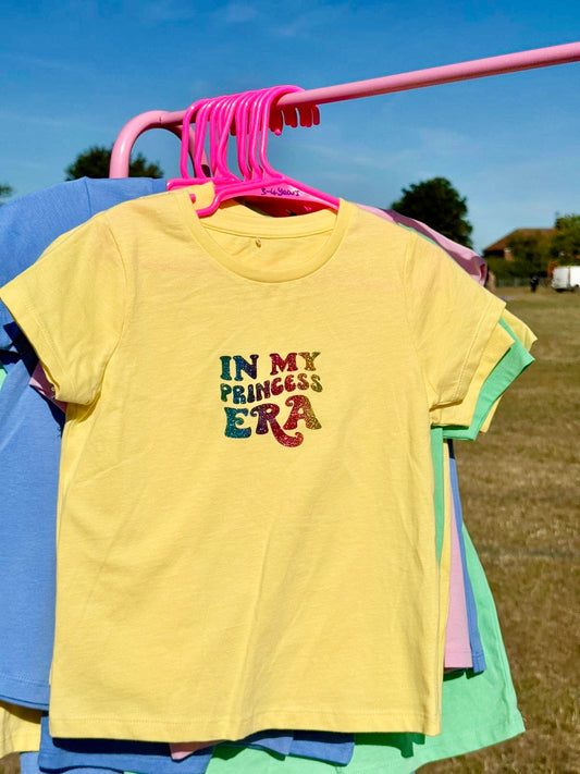 Yellow children’s t-shirt with ‘In My Princess Era’ in rainbow font, hanging on a pink hanger on a pink rail, photographed outdoors in the countryside.
