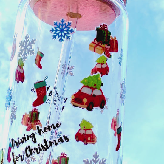 A reusable “Driving Home for Christmas” glass cup with bamboo lid and glass straw, decorated with red cars carrying presents, stockings, and snowflakes, held against a bright blue sky.