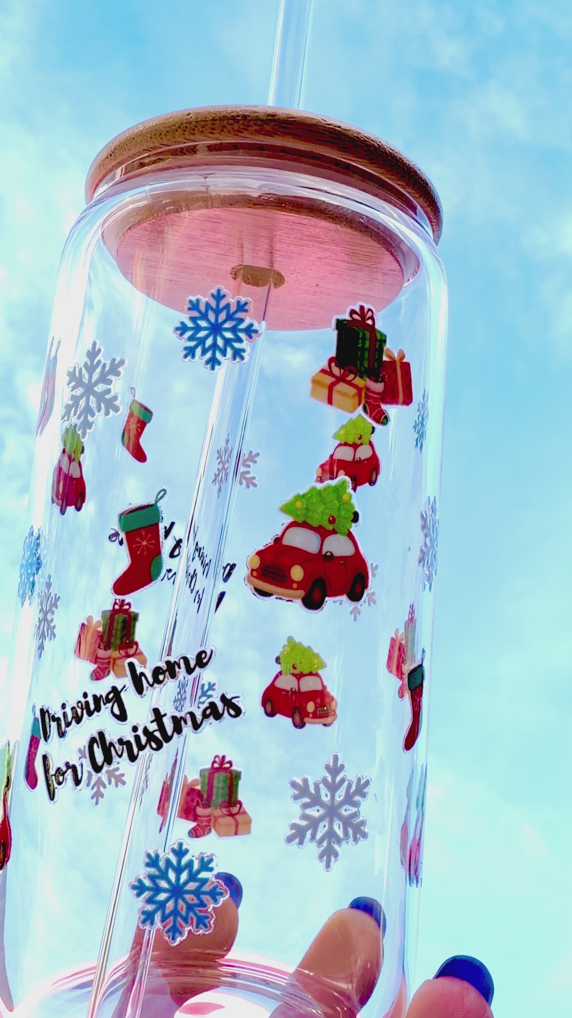 A reusable “Driving Home for Christmas” glass cup with bamboo lid and glass straw, decorated with red cars carrying presents, stockings, and snowflakes, held against a bright blue sky.