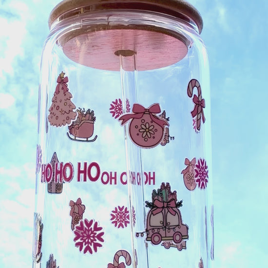 A reusable pink “HO HO HO” Christmas glass with bamboo lid and clear glass straw, decorated with candy canes, stockings, and gifts, held against a bright festive background and rotating