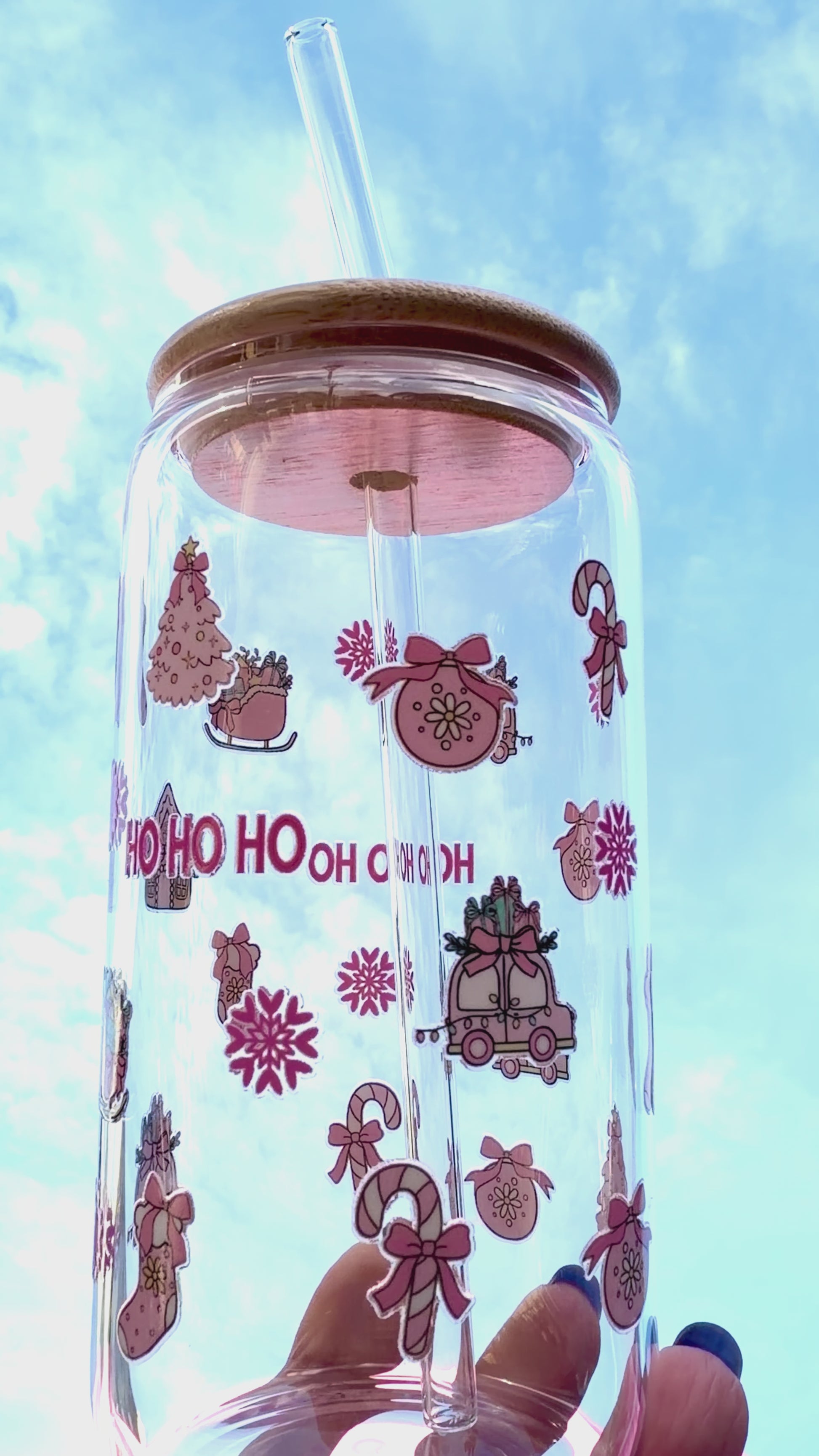 A reusable pink “HO HO HO” Christmas glass with bamboo lid and clear glass straw, decorated with candy canes, stockings, and gifts, held against a bright festive background and rotating