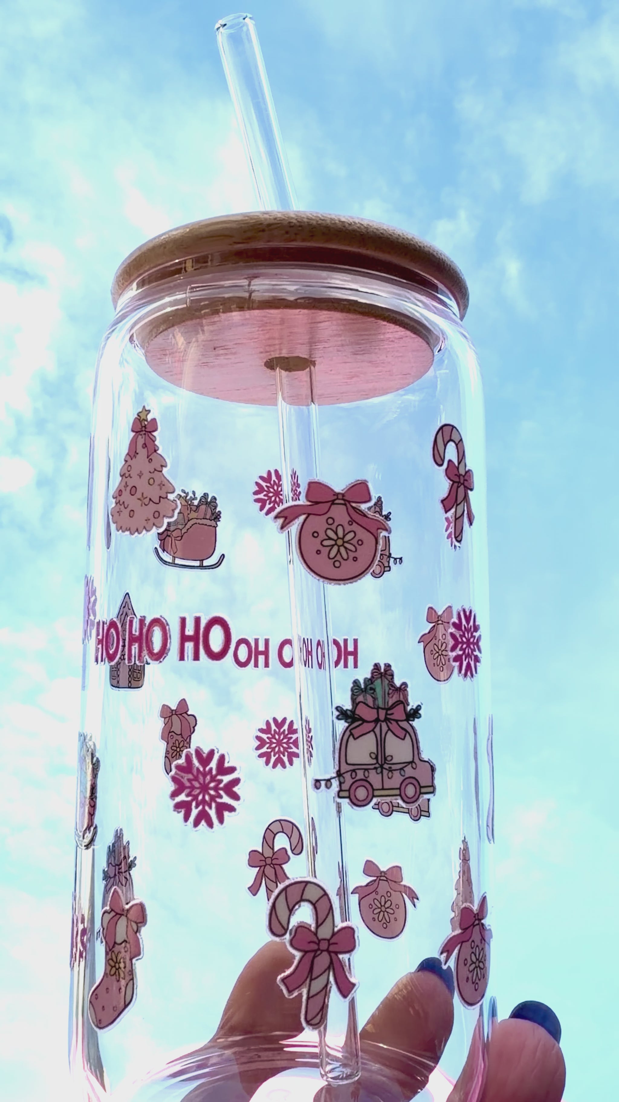 A reusable pink “HO HO HO” Christmas glass with bamboo lid and clear glass straw, decorated with candy canes, stockings, and gifts, held against a bright festive background and rotating