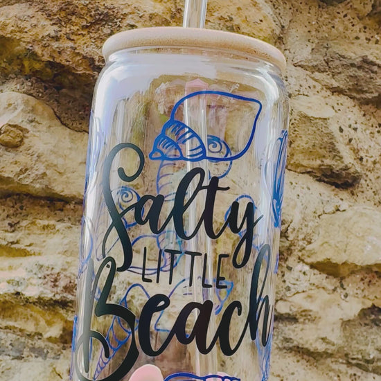 Video of a Glass Can Cup showing the black cursive font saying salty little beach and has blue sea shells all around the glass, with bamboo lid and glass straw