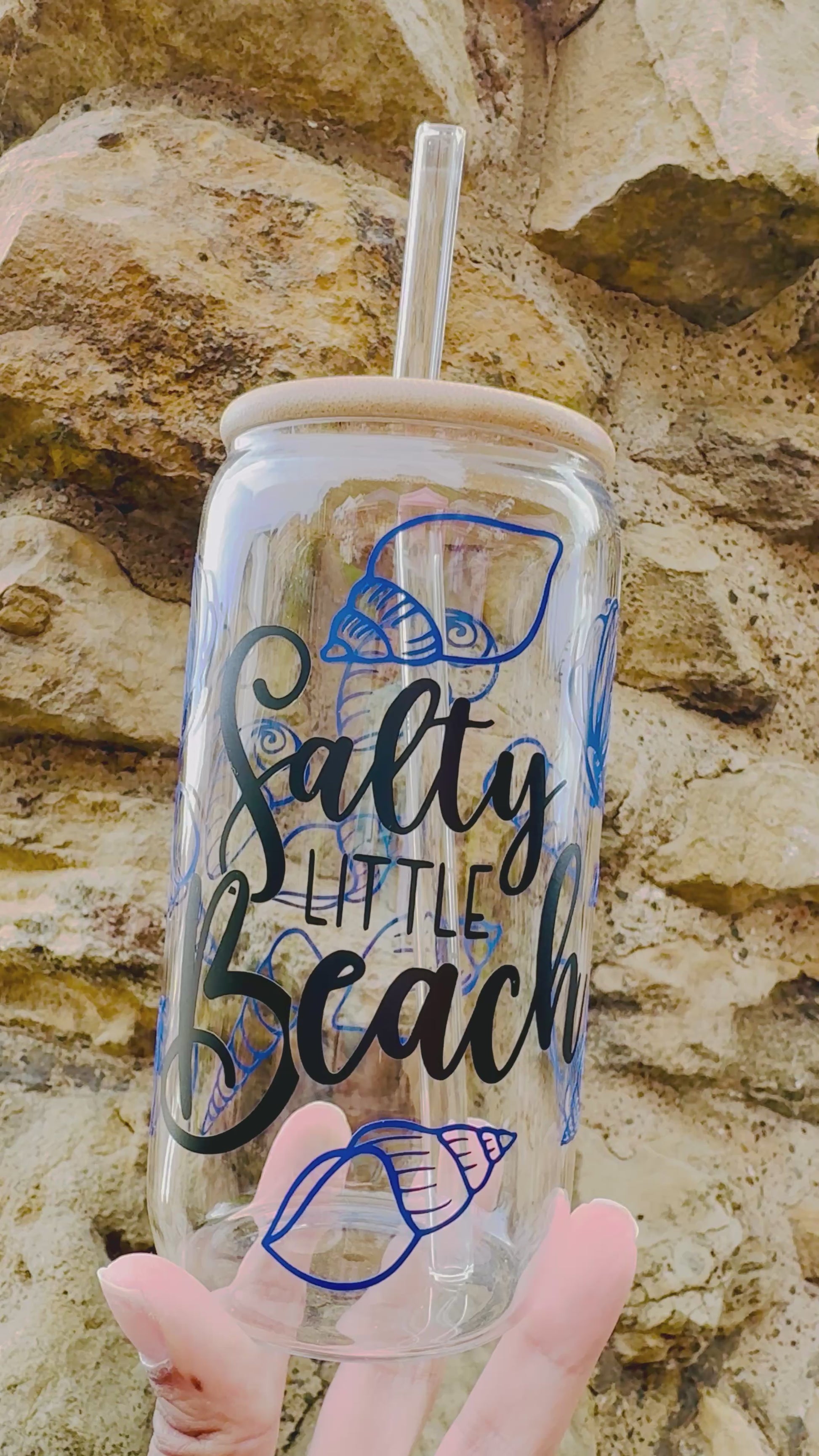 Video of a Glass Can Cup showing the black cursive font saying salty little beach and has blue sea shells all around the glass, with bamboo lid and glass straw